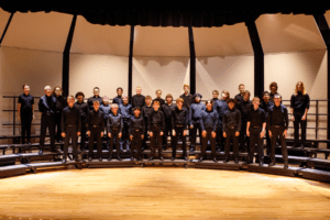 Male singers standing together in all black clothing and smiling